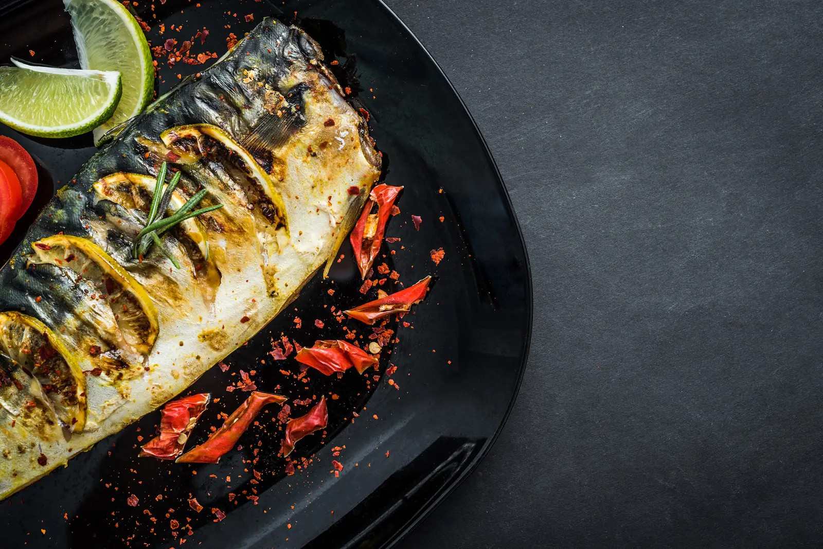 A whole grilled fish with lime, chili and herbs on a dark plate.
