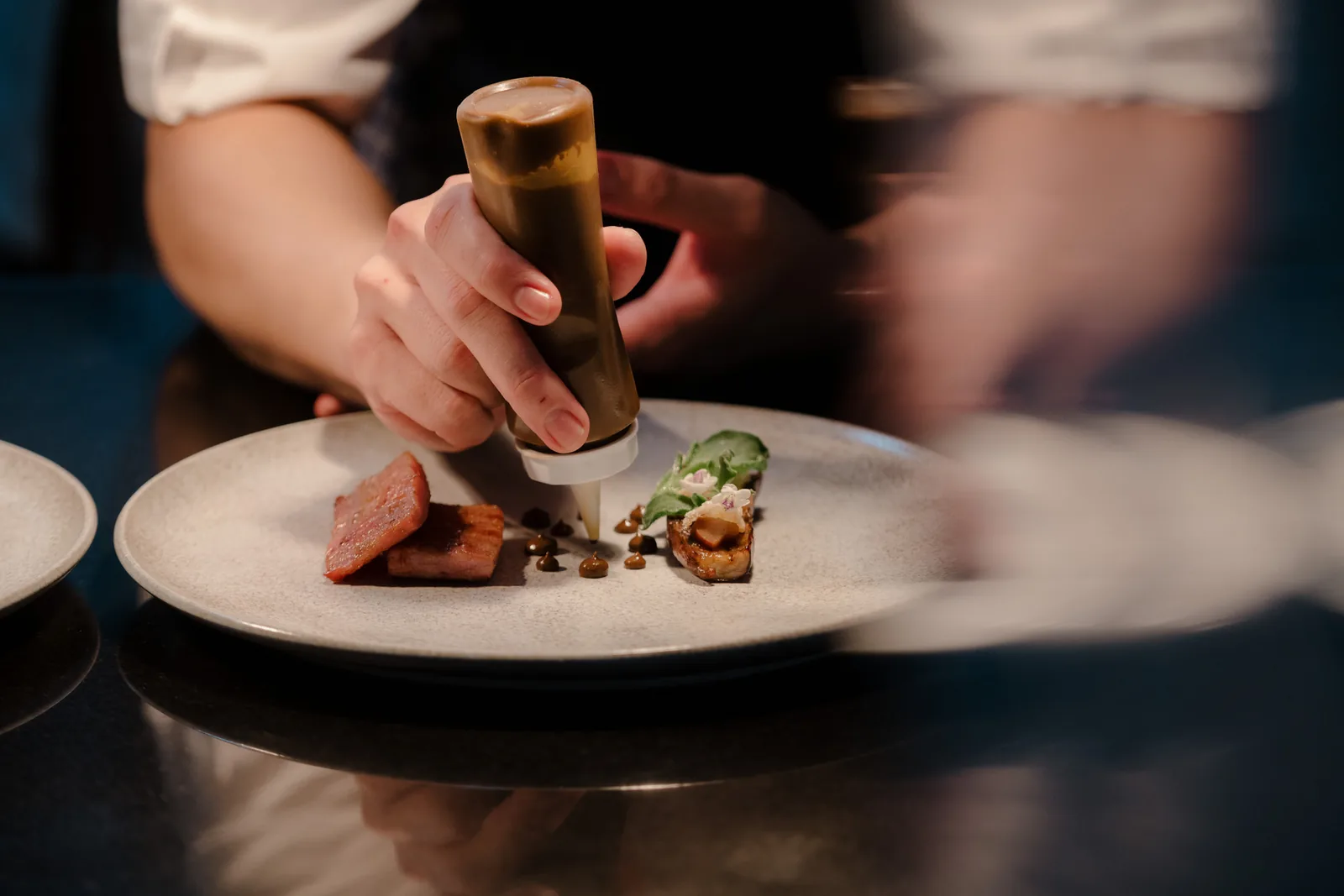 A cook's hands finishing a plated dish with a squeeze bottle on a dark pass.