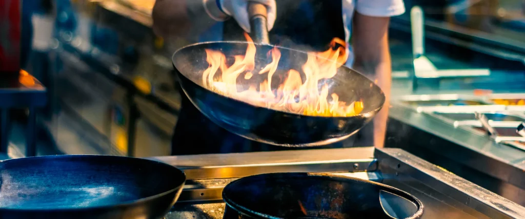 A cook tosses a flaming pan on a restaurant range during service.